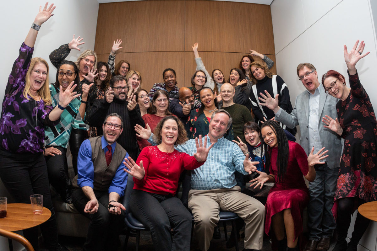 A group of about two dozen people in semi-formal business attire in a conference room set up with a holiday party atmosphere. Everyone is waving and smiling.