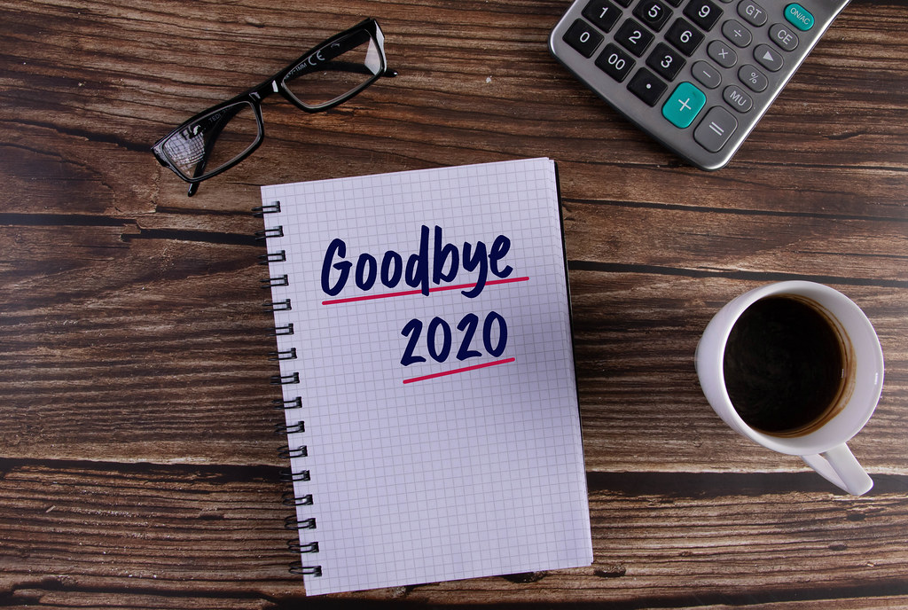 overhead photo of wooden desktop with calculator, half full coffee cup, glasses, and open notebook with "goodbye 2020" written in thick black letters with red underline